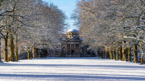 View of a snowy avenue of bare trees leading to a columned chapel covered in snow.